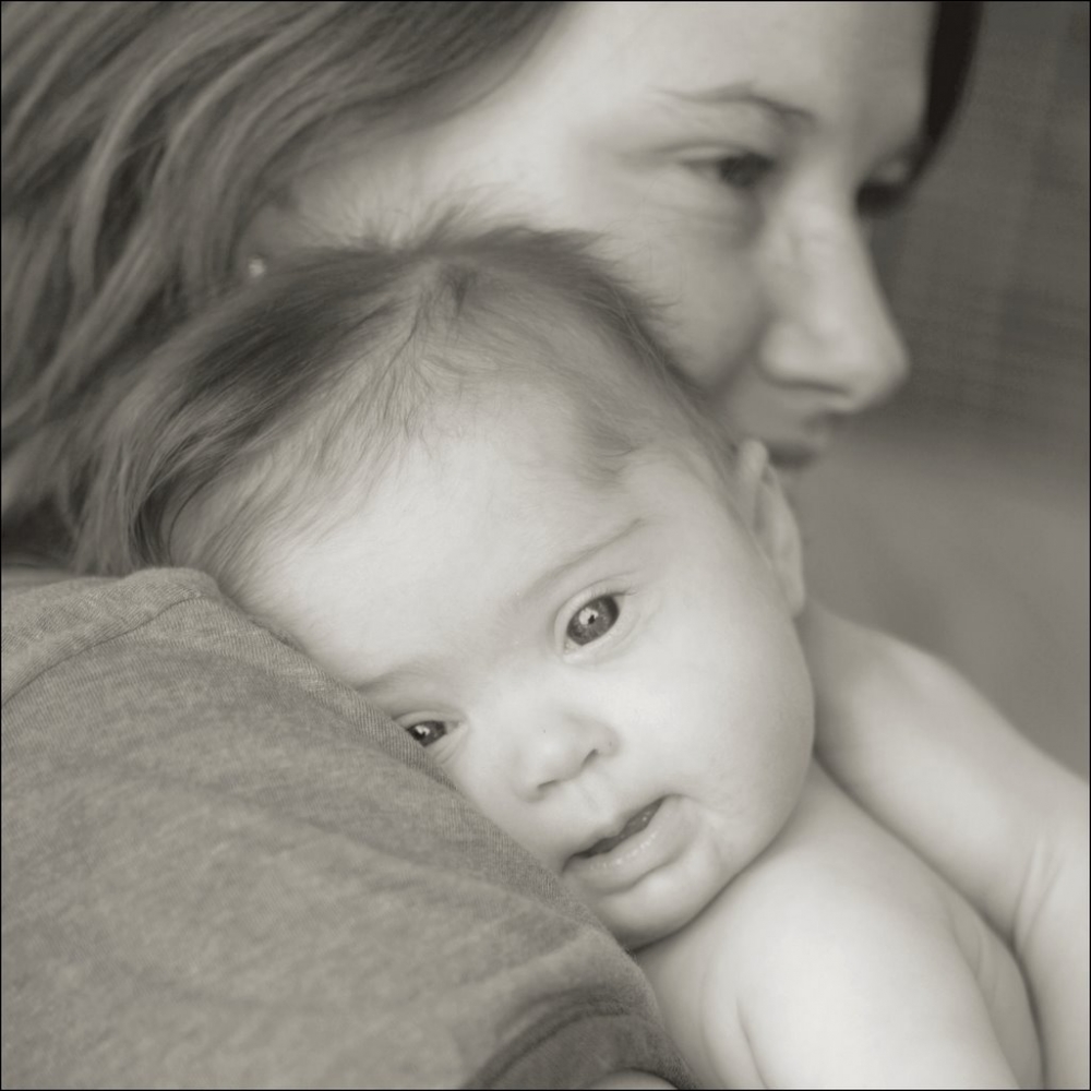 Pé de media (e descrición alternativa): Unha fotografía en branco e negro na que se apreza un bebé no colo da súa nai. O bebé mira cara a nós sorrinte e medio parece adiviñarse que ten o Síndrome de Down.