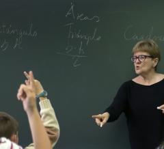 Mestra dando clase diante dun encerado, con alumnos de costas levantando a mán